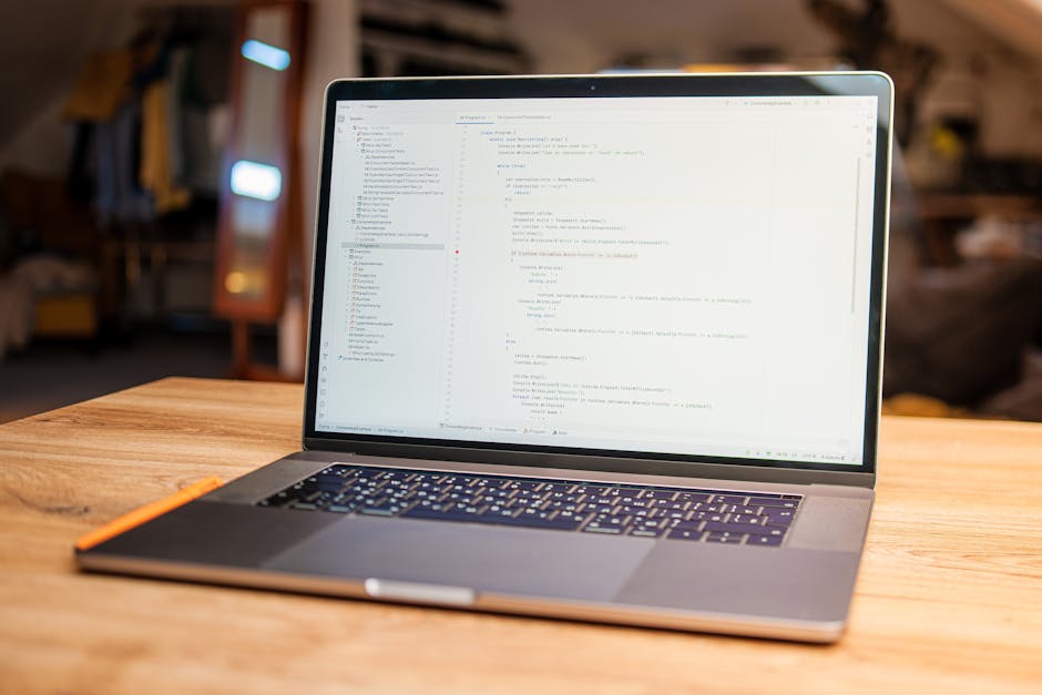A laptop placed on a wooden desk displaying lines of code on its screen, situated in a well-lit room with a blurred background that includes a bookshelf, a mirror, and a cozy living space. The laptop 